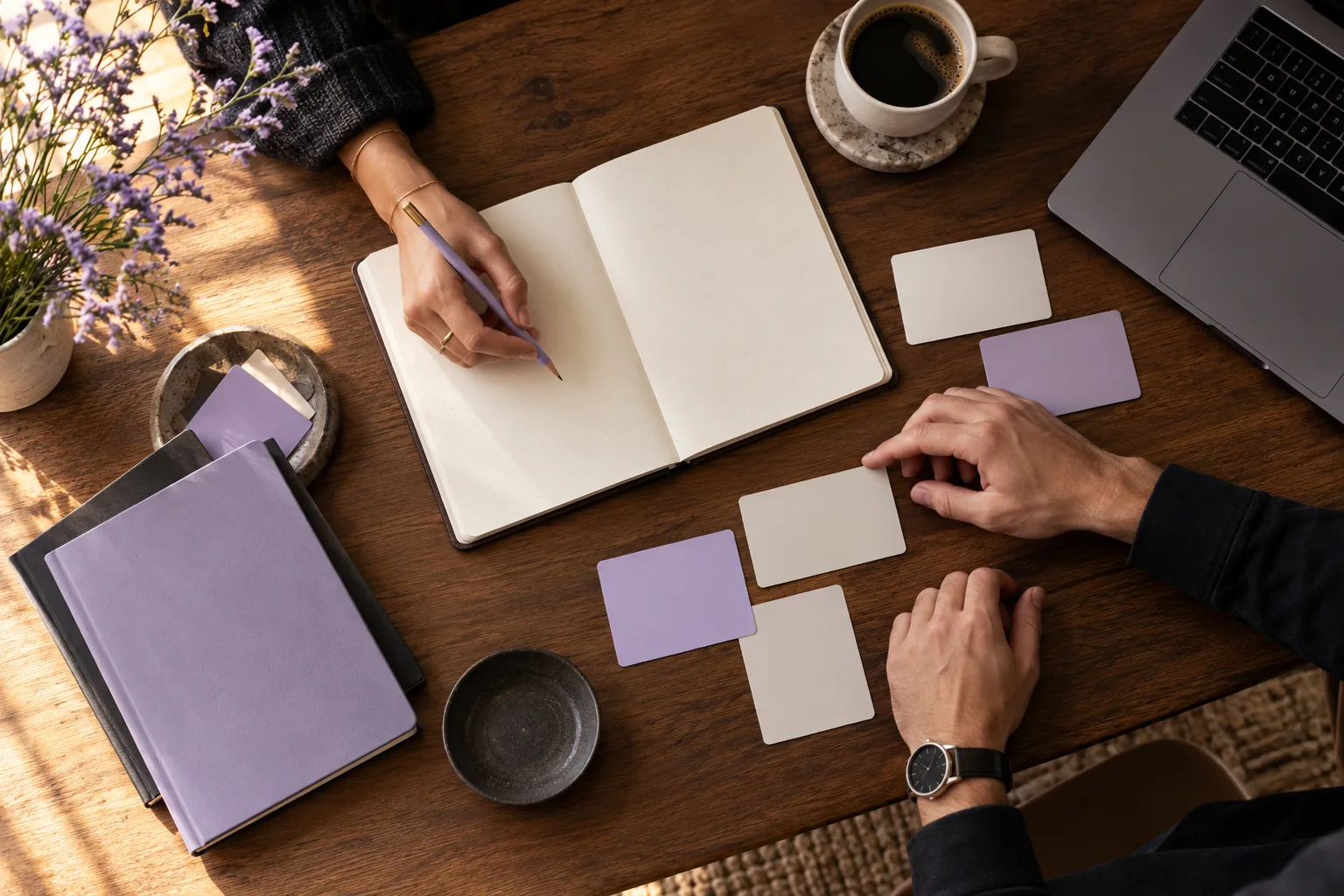 Hands arranging planning cards beside a notebook and laptop on a sunlit table